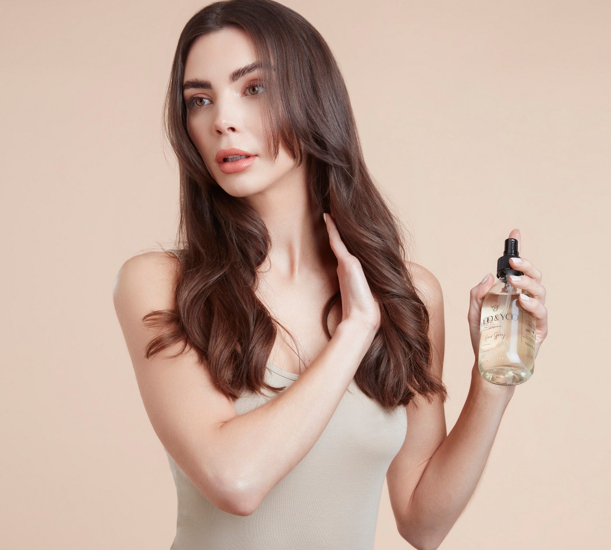 Woman with long, wavy brown hair holding a bottle of hair product against a beige background