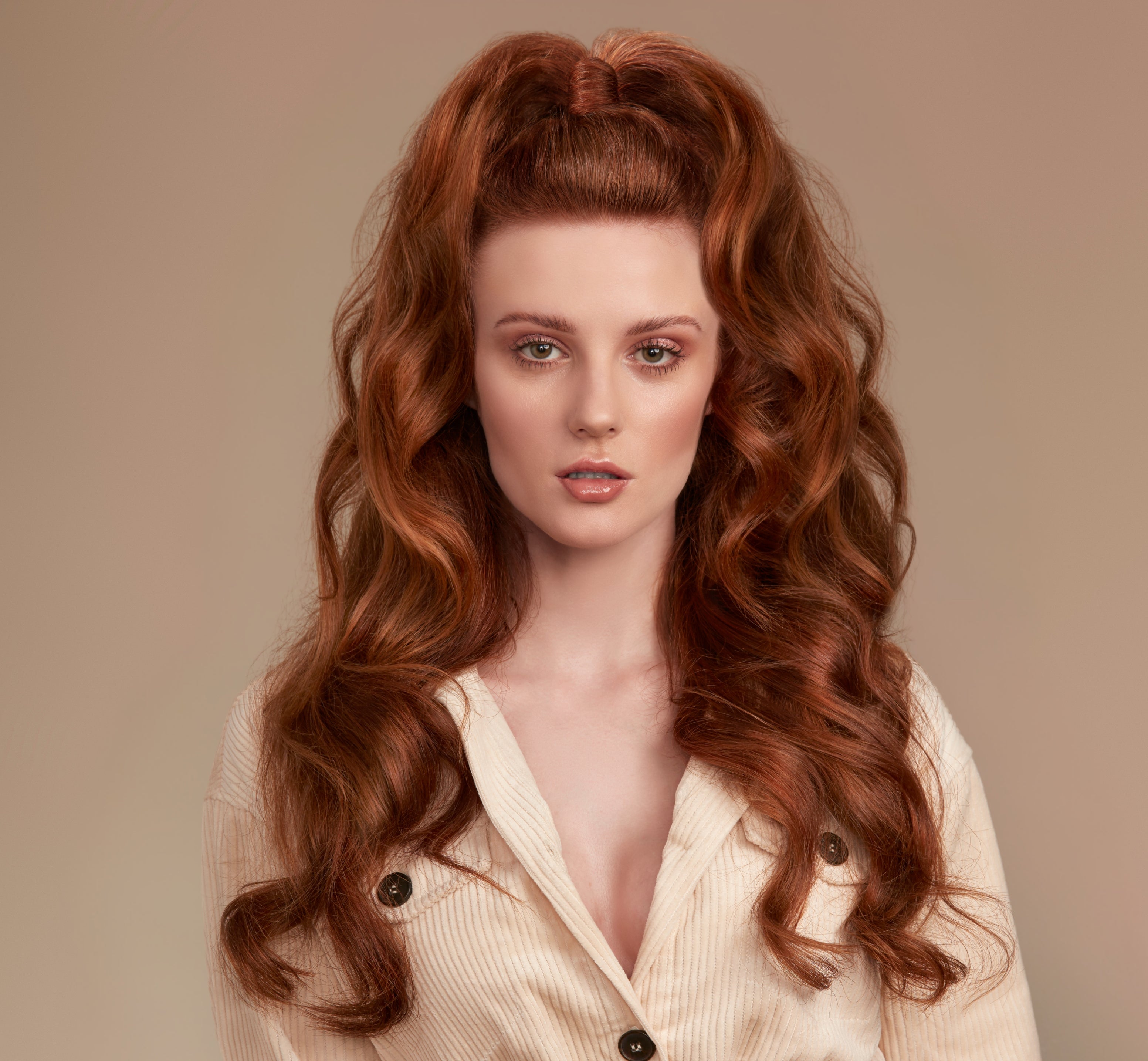 Woman with long, shiny wavy red hair half up wearing a light-colored jacket against a beige background looking at the camera. Her hair is very full with lots of volume but still looking glossy and shiny 
