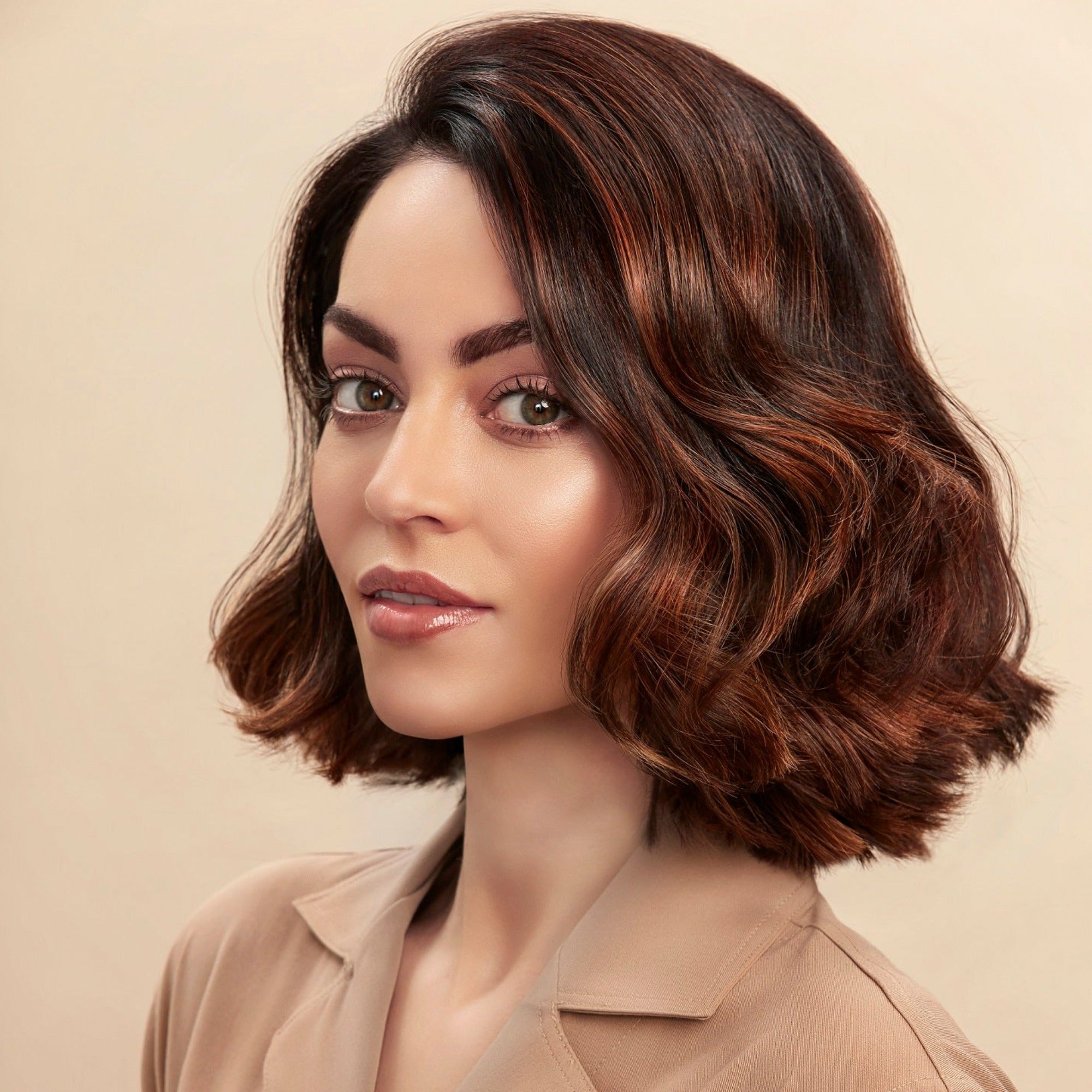 Female model with a wavy bob and brunette hair. She has super shiny hair with lots of volume. She is looking at the camera from a side angle and is wearing a neutral coloured shirt.