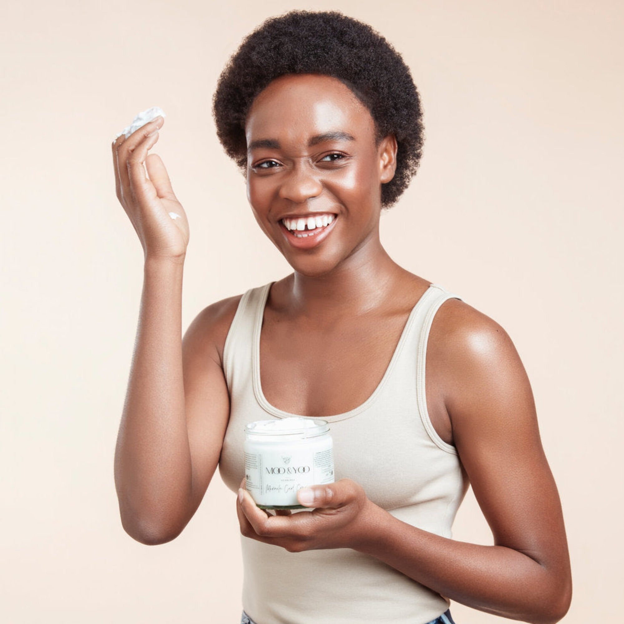Smiling female model laughing holding Moo and Yoo Miracle Curl Cream in one hand with a scoop of the product in her other hand held up to her hair. She has black textured natural hair and is wearing a neutral coloured vest top.