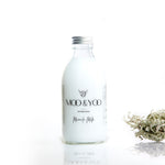 A glass bottle of Moo and Yoo Miracle Milk with an aluminium lid on a white background with a sprig of Icelandic moss placed to one side.