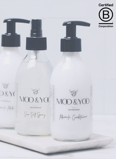 trio of products in glass bottles placed on stone slab diagonally. Front products is pour Miracle Conditioner, then Sea salt spray followed by our miracle milk. B corp logo is on the bottom right of the screen to represent Moo & Yoo being certified 