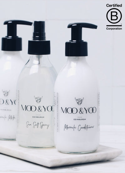 trio of products in glass bottles placed on stone slab diagonally. Front products is pour Miracle Conditioner, then Sea salt spray followed by our miracle milk. B corp logo is on the bottom right of the screen to represent Moo & Yoo being certified 