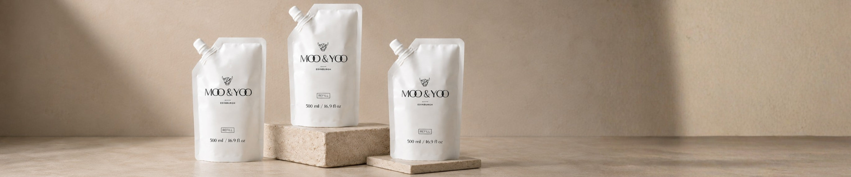 Three white Moo & Yoo refill pouches with a brand logo on a wooden surface and beige background