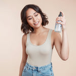 Woman with choppy short brunette holding a sea salt spray bottle up to her hair using product against a beige background