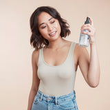Woman with choppy short brunette holding a sea salt spray bottle up to her hair using product against a beige background