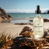 Moo & Yoo Sea Salt Spray bottle on a beach with shells and rocks.