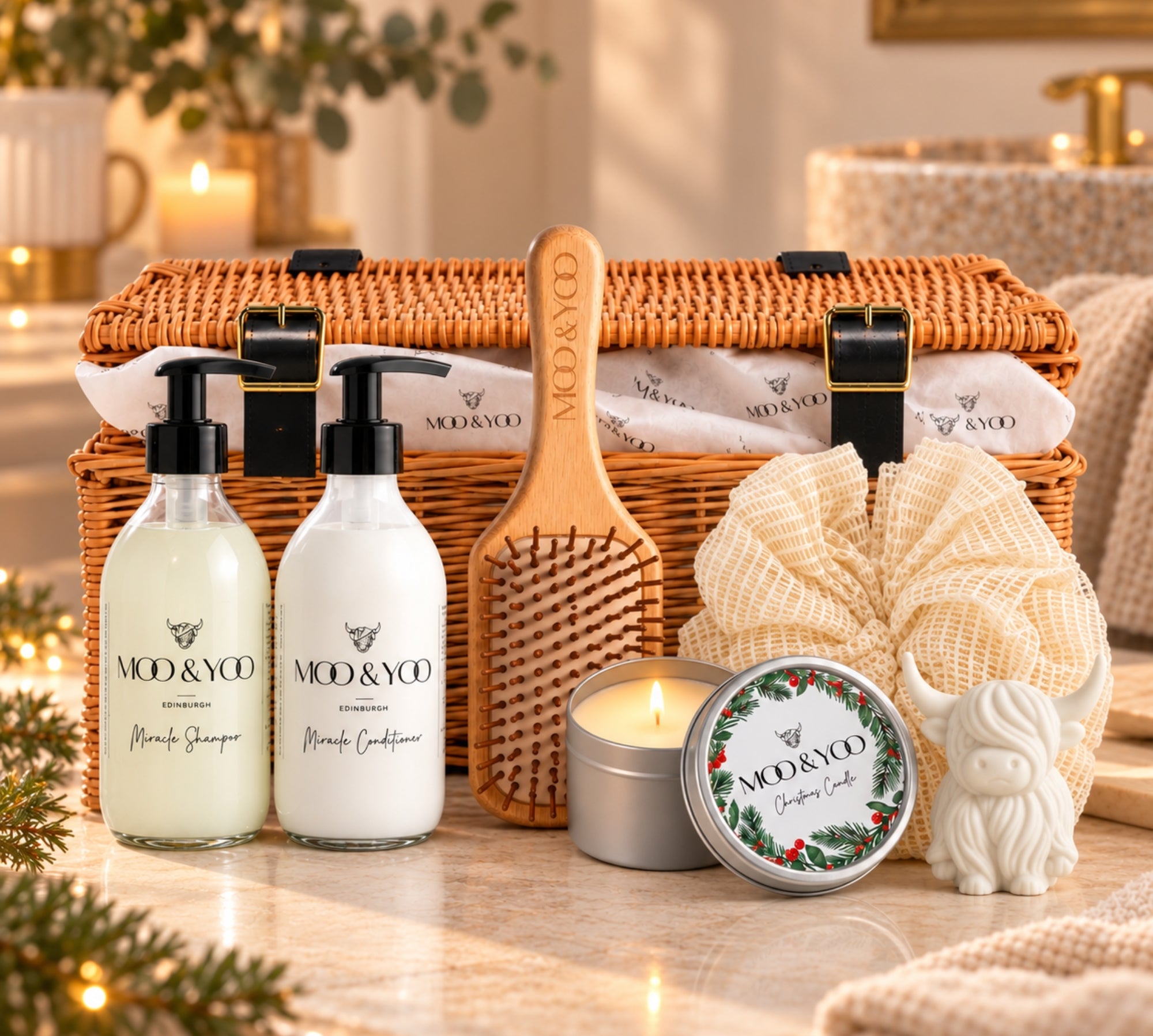 A spa luxury gift basket hamper set from MOO & YOO arranged in a wicker picnic basket on a marble surface. The set includes a bottle of miracle shampoo and a bottle of miracle conditioner, a wooden paddle hairbrush engraved with the brand name, a cream-colored hand towel, a small white decorative highland cow soap, a christmas candle tin.