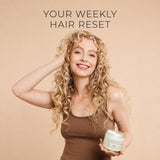 A smiling woman with long curly blonde hair holds a jar of moo and yoo miracle mask hair product against a warm beige background, with the text 'YOUR WEEKLY HAIR RESET' above her.