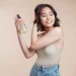 Woman holding a bottle of volumising spray mist spraying it inot her short brunette choppy style hair against a beige background