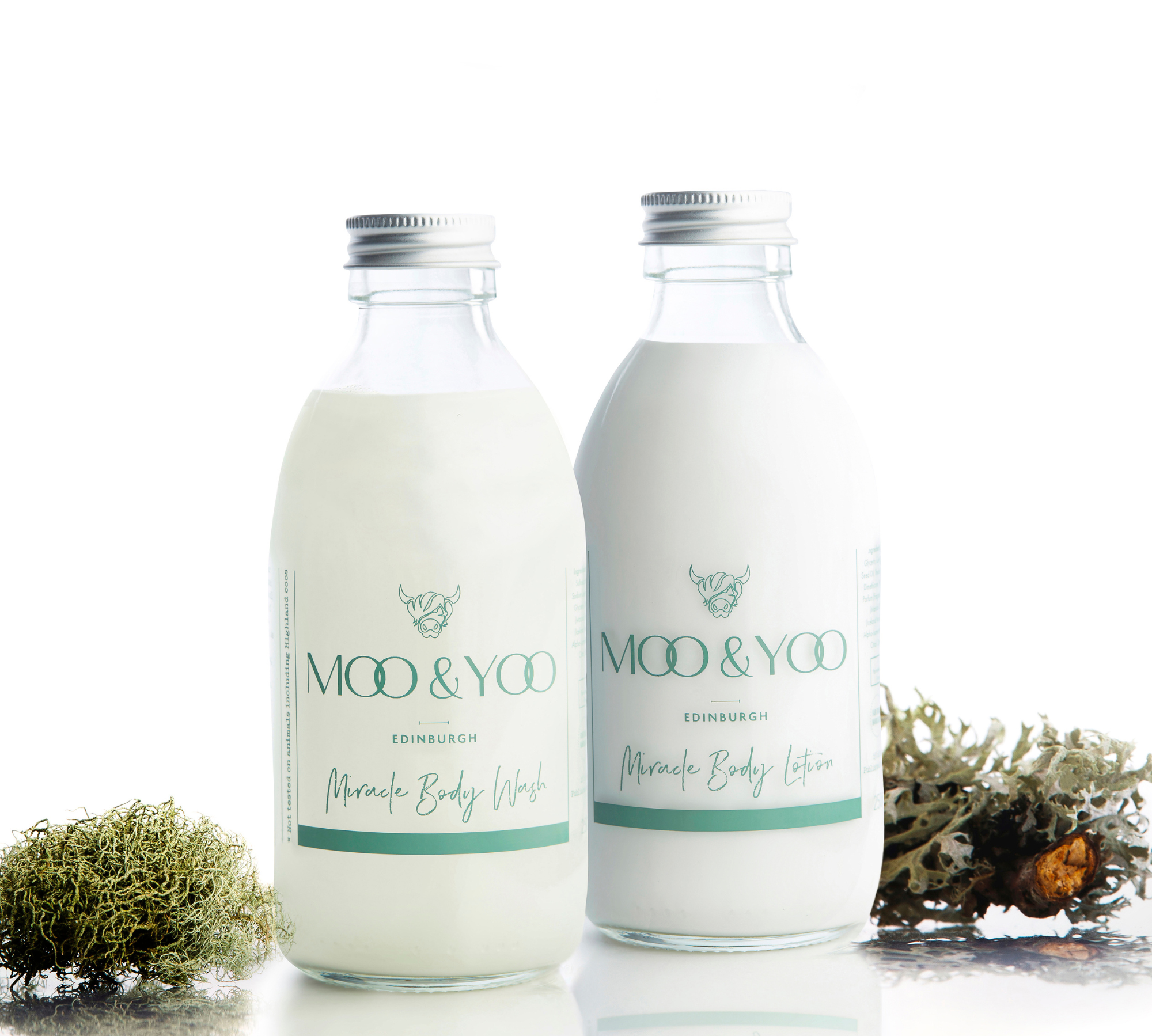 Two glass bottles containing Miracle Body Wash and Miracle Body Lotion sitting side by side on a white bacground with sprigs of moss on either side.