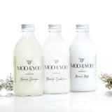 Three glass bottles of Moo and Yoo products sitting side by side. It is a Miracle Shampoo, Conditioner and a Miracle Milk. All have aluminium lids.  They are on a white background with a sprig of Icelandic moss placed to each side.