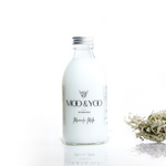 A glass bottle of Moo and Yoo Miracle Milk with an aluminium lid on a white background with a sprig of Icelandic moss placed to one side.