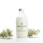 A glass bottle of Moo and Yoo Miracle shampoo with an  aluminium lid on a white background with a sprig of icelandic moss placed to each side.