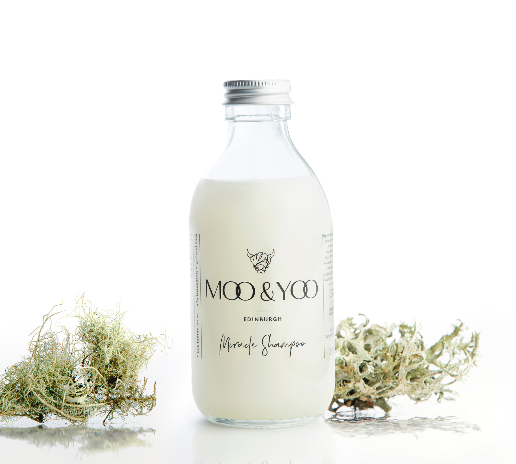 A glass bottle of Moo and Yoo Miracle shampoo with an  aluminium lid on a white background with a sprig of icelandic moss placed to each side.