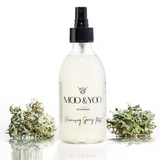 A glass jar of Moo and Yoo Volumising Spray Mist with a spray lid on a white background with a sprig of Icelandic moss placed to each side.